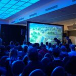 A seated audience in a conference room watching gameplay on a large projection screen under blue stage lighting