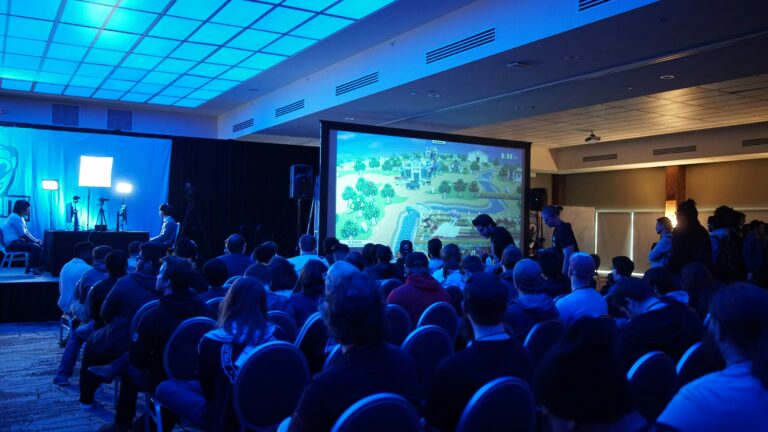 A seated audience in a conference room watching gameplay on a large projection screen under blue stage lighting